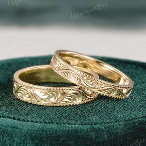 Puede incluir: Dos anillos de oro con un intrincado patrón de volutas grabado a lo largo de la banda exterior. Cada anillo tiene un acabado pulido con un motivo floral detallado y un borde con cuentas, mostrando un diseño clásico en metal de oro amarillo.