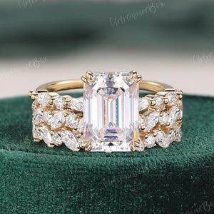 May include: A gold engagement ring set with a large emerald cut center stone and a matching band with smaller round stones. The ring is on a green velvet box.