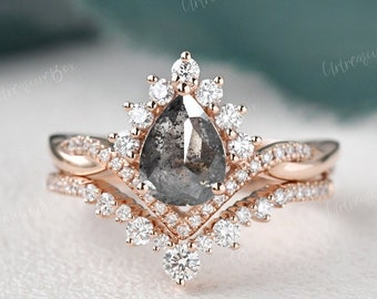 Puede incluir: Un conjunto de anillos de oro rosa con un diamante central en forma de pera tipo sal y pimienta, rodeado por un halo de diamantes redondos de talla brillante. El anillo tiene un diseño trenzado y se acompaña de una alianza en forma de V.