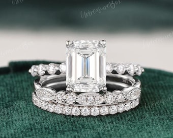 May include: A silver-toned engagement ring set featuring a large emerald-cut center stone, flanked by two matching bands. The bands are adorned with small, round, clear stones in a pavé setting, creating a layered, sparkling appearance.