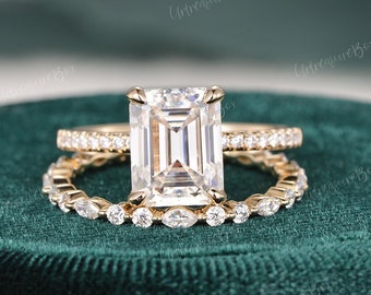 May include: A gold engagement ring set with a large emerald cut center stone and a matching band with smaller round stones. The ring is on a green velvet box.