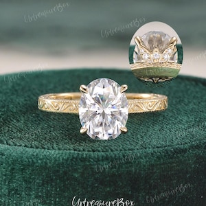 May include: A gold engagement ring with an oval-cut diamond. The ring features an intricate filigree design on the band and a detailed setting. The diamond is the focal point, reflecting light. The ring is displayed on a green velvet surface.