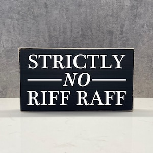 Peut inclure: Panneau en bois noir avec l'inscription blanche "STRICTLY - NO - RIFF RAFF". Le panneau rectangulaire est posé sur une surface blanche, devant un fond gris. Le texte est centré et en gras, avec une police sans empattement.