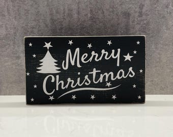 Può includere: Un cartello rettangolare in legno nero con testo bianco che recita "Merry Christmas". Sono presenti anche un albero di Natale bianco e delle stelle. Il cartello è una decorazione festiva.