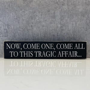 May include: A black wooden sign with white text that reads "NOW, COME ONE, COME ALL TO THIS TRAGIC AFFAIR...". The sign is rectangular and sits on a white surface.