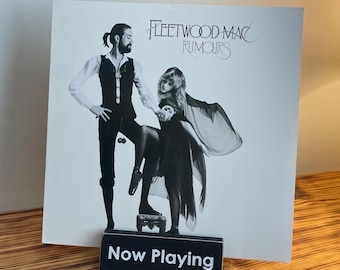 May include: A Fleetwood Mac "Rumours" album cover featuring a black and white image of a man and woman. The album is displayed on a wooden surface with a "Now Playing" sign in front. The album cover has the band's name and album title.