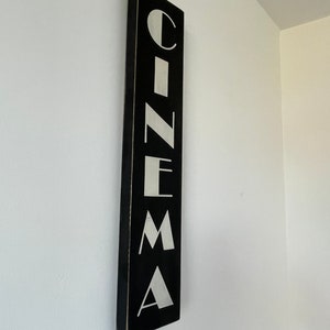 以下が含まれることがあります： 白い壁に掛けられた、黒い木製の看板に白い文字で「CINEMA」と書かれている。