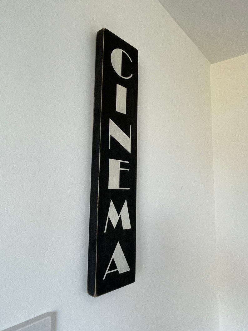 以下が含まれることがあります： 白い壁に掛けられた、黒い木製の看板に白い文字で「CINEMA」と書かれている。