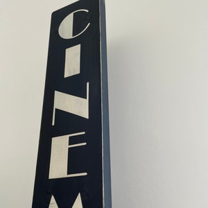 以下が含まれることがあります： 白い文字で「CINEMA」と書いてある黒い木製の看板。看板は縦型で、使い込まれたような風合いがある。