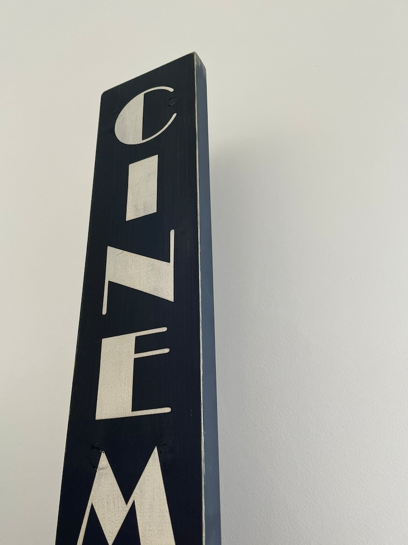 以下が含まれることがあります： 白い文字で「CINEMA」と書いてある黒い木製の看板。看板は縦型で、使い込まれたような風合いがある。