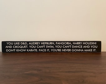 May include: Black wooden sign with white text that reads: "YOU LIKE D&D, AUDREY HEPBURN, FANGORIA, HARRY HOUDINI AND CROQUET. YOU CAN'T SWIM, YOU CAN'T DANCE AND YOU DON'T KNOW KARATE. FACE IT, YOU'RE NEVER GONNA MAKE IT."
