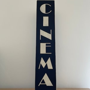 以下が含まれることがあります： 濃い青色の背景に白い文字で「CINEMA」と書かれた縦型の木製看板です。