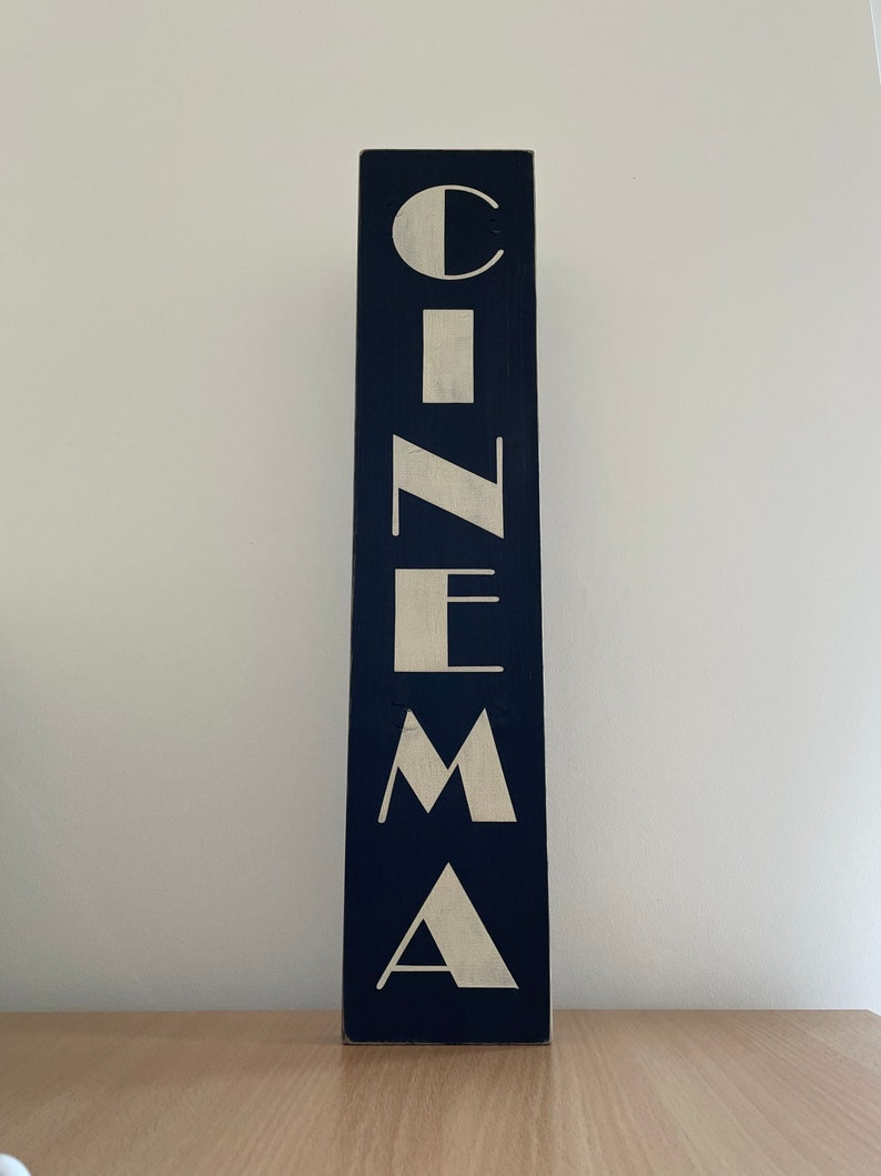 以下が含まれることがあります： 濃い青色の背景に白い文字で「CINEMA」と書かれた縦型の木製看板です。