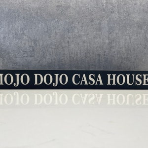 Puede incluir: Un letrero rectangular de madera negra con las palabras "MOJO DOJO CASA HOUSE" en letras blancas. El letrero está sobre una superficie blanca, con un fondo gris texturizado. Se ve el reflejo del letrero.