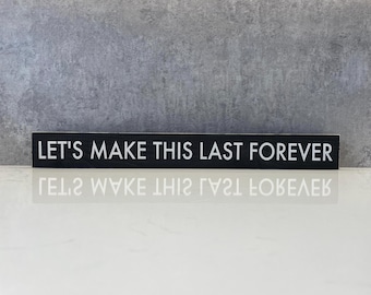 以下が含まれることがあります： 「LET'S MAKE THIS LAST FOREVER」の白い文字が書かれた長方形の黒い木製の看板。白い表面に置かれ、背景はグレーです。