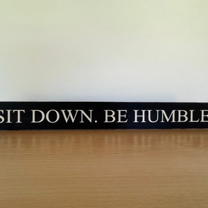 Könnte beinhalten: Schwarzes Holzschild mit weißem Text, der "SIT DOWN. BE HUMBLE." lautet.
