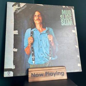 Può includere: È esposta una copertina di un album in vinile vintage di "Mud Slide Slim" di James Taylor. La copertina dell'album presenta un ritratto di James Taylor. Un cartello di legno con la scritta "Now Playing" è di fronte all'album.