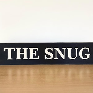 Peut inclure: Une pancarte en bois noir avec l'inscription "THE SNUG" en lettres blanches.