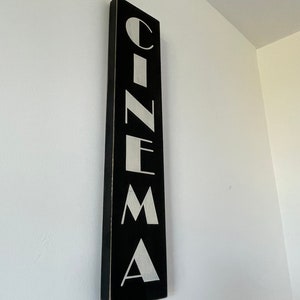 以下が含まれることがあります： 白い文字で「CINEMA」と書かれた黒い木製の看板。文字はスタイリッシュで、ビンテージな雰囲気です。