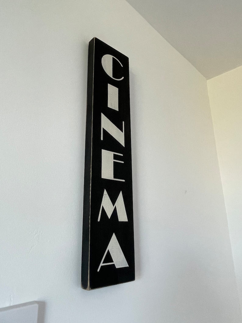 以下が含まれることがあります： 白い文字で「CINEMA」と書かれた黒い木製の看板。文字はスタイリッシュで、ビンテージな雰囲気です。
