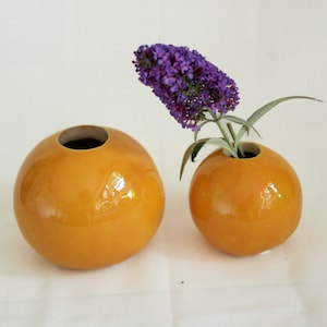 Könnte beinhalten: Zwei runde Keramikvasen in leuchtend gelber Farbe. Die kleinere Vase hält einen Strauß lila Blumen.