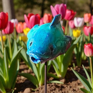 Könnte beinhalten: Ein leuchtend blau-grüner Keramik-Fisch-Gartenstecker mit offenem Maul und dunklen Augen. Der Fisch ist vor einem Hintergrund aus bunten Tulpen platziert. Der Fisch ist auf einem dünnen Metallstab montiert.
