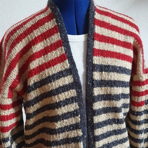 Könnte beinhalten: Ein gestreifter Cardigan in Rot, Beige und Marineblau. Der offene Strickjacke hat horizontale Streifen und einen Schalkragen. Das Strickwaren wird auf einer Büste präsentiert, um die Textur und das Design hervorzuheben. Ein gemütlicher und stilvoller Oberbekleidungsartikel.