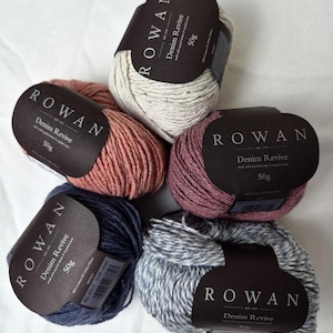 Könnte beinhalten: Fünf Knäuel Rowan Denim Revive Garn in verschiedenen Farben, darunter Rosa, Blau und Grau. Jedes Knäuel ist mit dem Rowan-Logo und dem Text "Denim Revive 50g" versehen. Das Garn wird aus recycelten Baumwollfasern hergestellt.