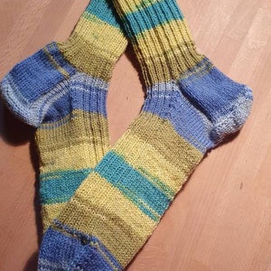 May include: A pair of knitted socks in blue, yellow, and green stripes. The socks are folded in half and laid flat on a wooden surface.