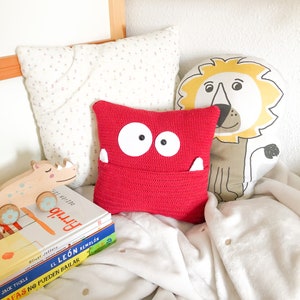 Puede incluir: Una almohada roja de crochet con cara de monstruo y un bolsillo. La almohada está sobre una manta blanca con una almohada de león amarilla y una pila de libros infantiles.