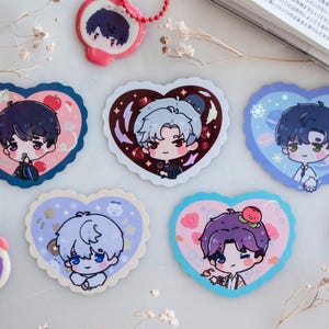 Op de afbeelding: Een verzameling hartvormige stickers en sleutelhangers met illustraties van personages in anime-stijl. De stickers en sleutelhangers hebben geschulpte randen en zijn versierd met verschillende kleuren en ontwerpen. Sommige hebben sleutelhangers.