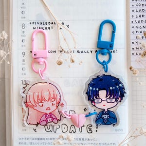 May include: Two anime-style acrylic keychains with cartoon characters. One keychain has a pink clasp and a pink-haired character. The other has a blue clasp and a blue-haired character. The word "UPDATE" is visible.