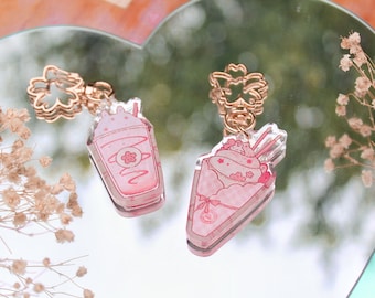 May include: Two pink and white acrylic charms on gold chains. One charm is a pink milkshake with a cherry on top. The other charm is a pink crepe with whipped cream and a cherry on top.