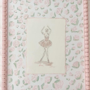May include: A framed artwork featuring a ballerina sketch in a pink tutu. The frame is light pink with a beaded edge. The background has a floral pattern of pink roses and green leaves on a white background.