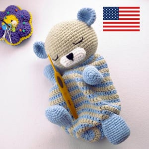 Puede incluir: Un oso de peluche tejido a crochet con una manta a rayas azul y beige. El oso tiene los ojos cerrados y una nariz blanca. El juguete está acostado sobre una superficie blanca.