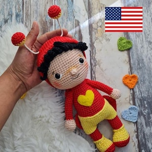 Puede incluir: Muñeca de ganchillo hecha a mano en rojo, amarillo y beige. La muñeca tiene un sombrero rojo con antenas, un corazón amarillo en el pecho y pantalones cortos amarillos. La muñeca se sostiene en una mano, con corazones de ganchillo y una bandera estadounidense en el fondo.