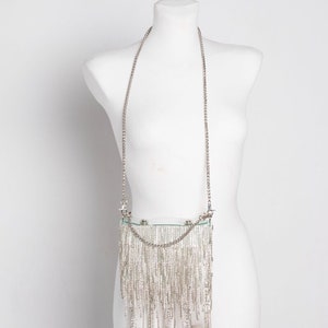 May include: A silver-colored, rectangular handbag with a long chain strap. The bag is adorned with numerous dangling silver strands, creating a fringe effect. The bag is displayed on a white mannequin.