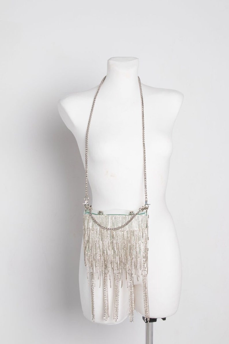 May include: A silver-colored, rectangular handbag with a long chain strap. The bag is adorned with numerous dangling silver strands, creating a fringe effect. The bag is displayed on a white mannequin.