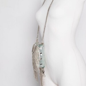 May include: A clear, rectangular handbag with a silver chain strap and dangling silver fringe. The bag has silver studs and a silver chain closure. The bag is displayed on a white mannequin.