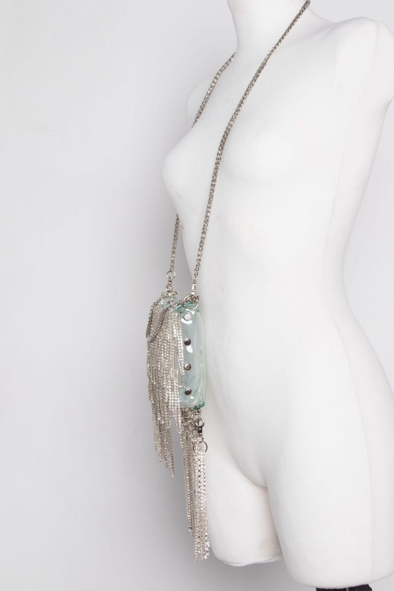 May include: A clear, rectangular handbag with a silver chain strap and dangling silver fringe. The bag has silver studs and a silver chain closure. The bag is displayed on a white mannequin.