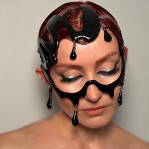 May include: A black, abstract headpiece and face mask with a glossy finish. The headpiece curves over the forehead and around the ears, while the mask covers the eyes and nose. Black teardrop-shaped pendants hang from the headpiece and mask.