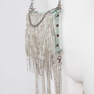 May include: A clear, rectangular handbag with a silver chain strap and a cascade of dangling silver rhinestones. The bag has a transparent body with silver hardware and is attached to a silver chain strap.