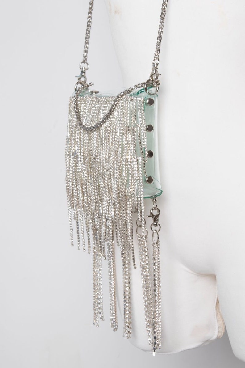 May include: A clear, rectangular handbag with a silver chain strap and a cascade of dangling silver rhinestones. The bag has a transparent body with silver hardware and is attached to a silver chain strap.
