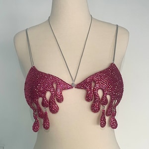 May include: A pink rhinestone bra with a dripping design. The bra is made of silver chain and rhinestones.