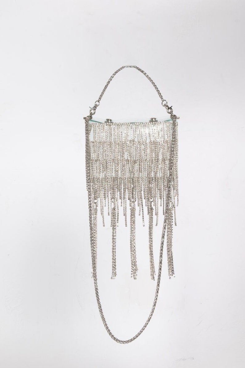 May include: A silver-toned handbag with a rectangular top and a chain handle. The bag is adorned with multiple strands of dangling silver chains, creating a fringe effect. The bag has a chain strap for shoulder or hand carry.