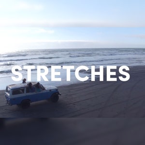 May include: A blue and white vintage SUV drives on a dark sandy beach with the word "STRETCHES" in white text across the image. The ocean and sky are visible in the background.