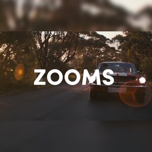 May include: A vintage red car with white racing stripes drives down a tree-lined road. The word "ZOOMS" is displayed in large white letters across the image. The car's headlights are on, and the license plate is visible.