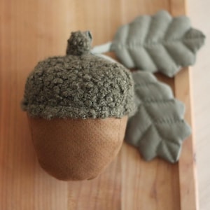 May include: A brown and green felt acorn with a fuzzy top, sitting on a wooden surface with two green felt leaves.