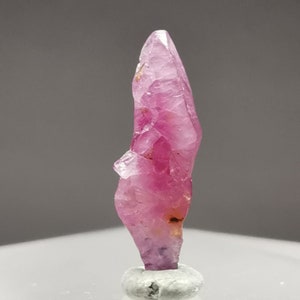 Puede incluir: Un cristal de rubí rosa vibrante con una punta puntiaguda y una superficie facetada. El cristal se muestra en un soporte blanco.