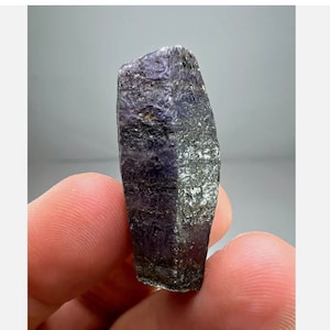 May include: A dark purple and gray crystal with a rough, faceted surface. The crystal is held in a hand.